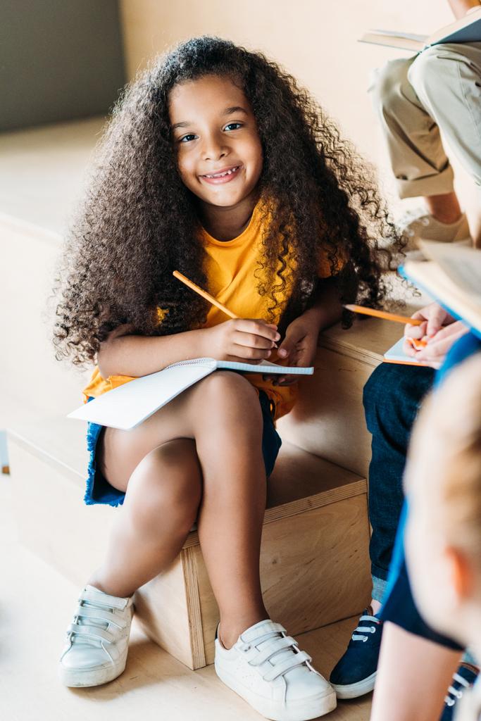 adorable smiling african american schoolgirl writing in notebook and looking at camera - Photo, Image