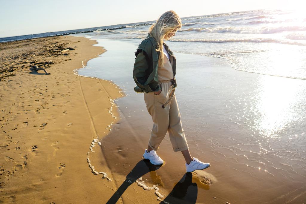 вид сбоку на молодую женщину в стильной одежде, идущую по берегу моря в летний день
 - Фото, изображение