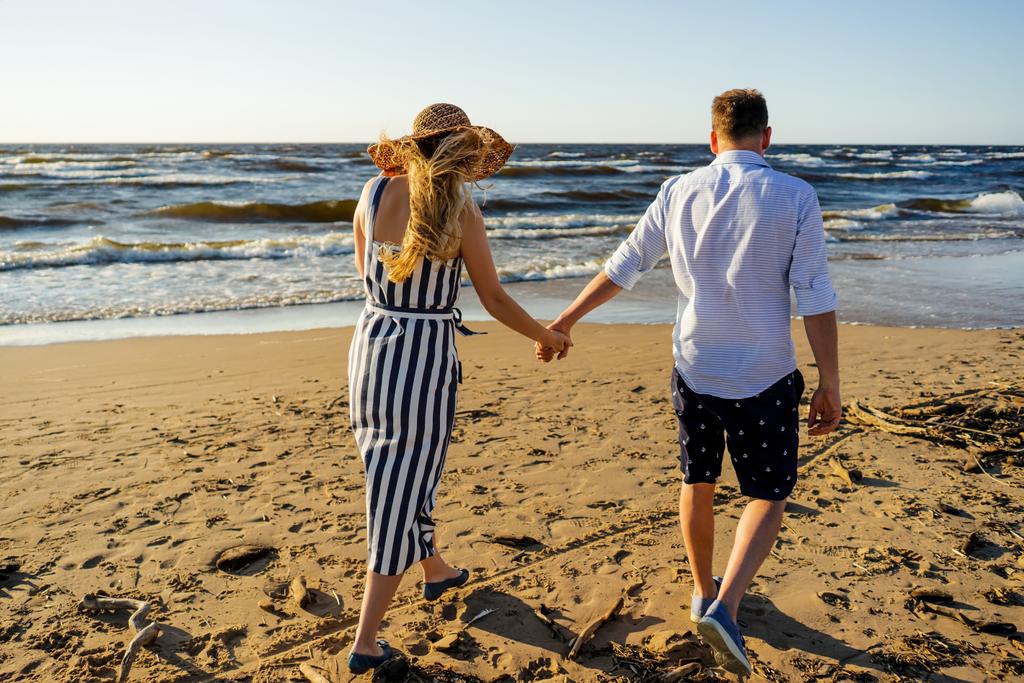 vista trasera de pareja cariñosa cogida de la mano y caminando en la playa de arena en el día de verano
 - Foto, imagen