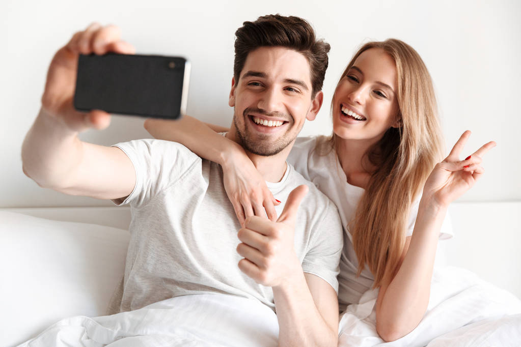 Image of young cheerful loving couple lies in bed take a selfie by smartphone looking aside showing peace gesture. - Photo, Image