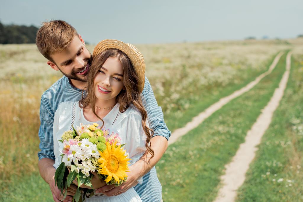 портрет усміхненого чоловіка, що обіймає дівчину з букетом диких квітів на літньому полі
 - Фото, зображення