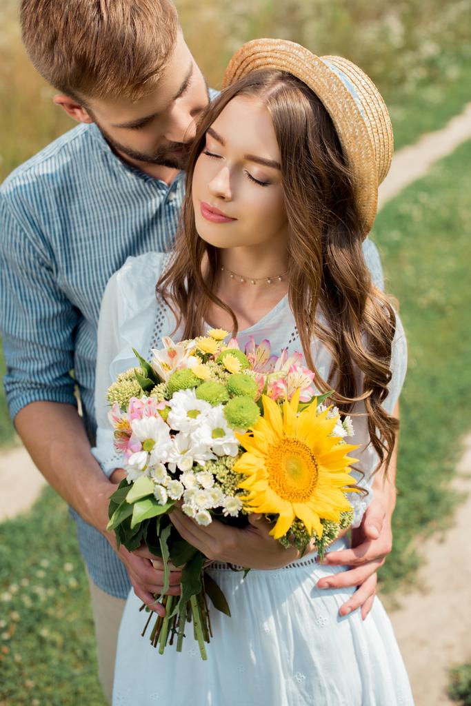 tedere man knuffelen vriendin met bouquet van veldbloemen in zomer veld - Foto, afbeelding