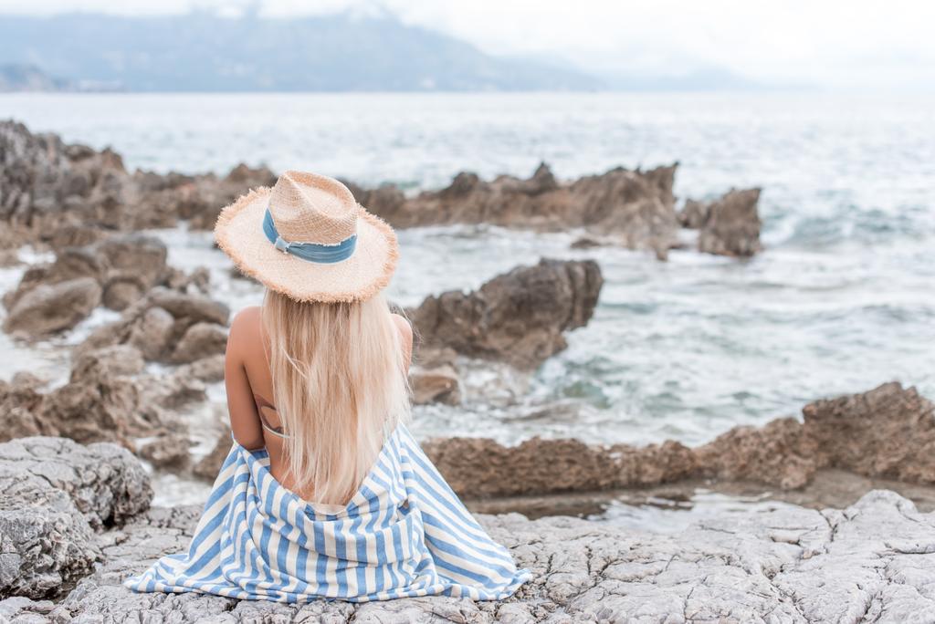 Achteraanzicht van meisje in stro hoed zittend op rotsachtige strand in montenegro - Foto, afbeelding
