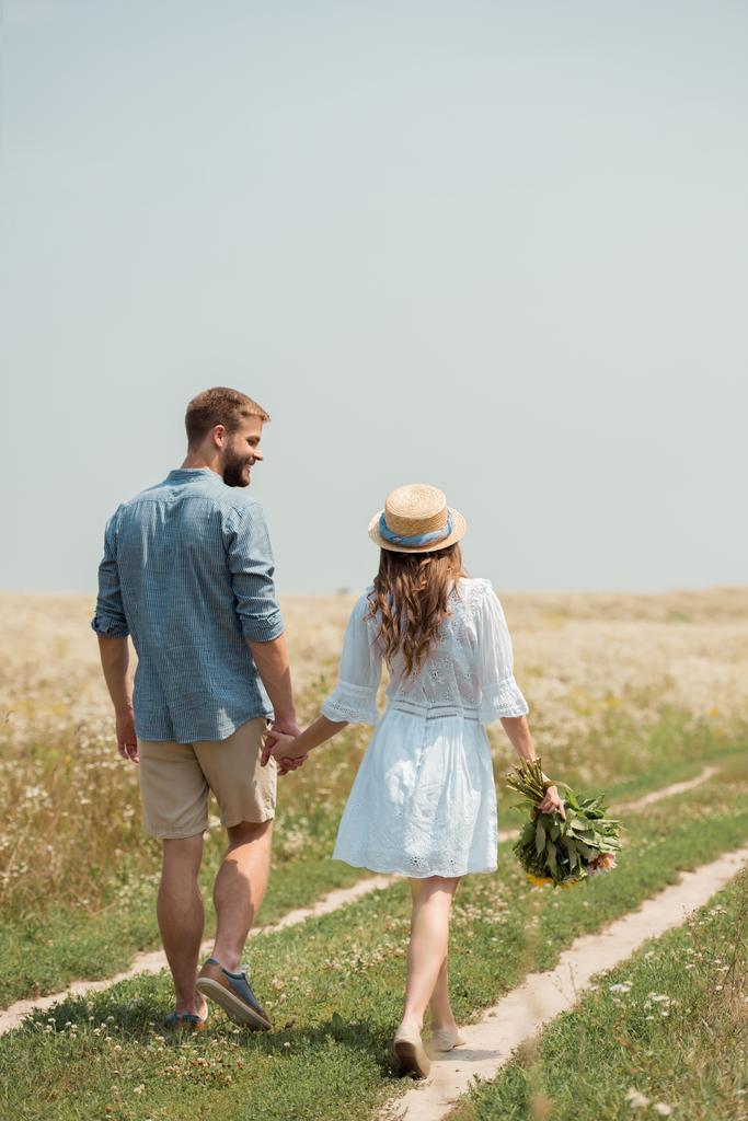 vista posterior de la mujer en vestido blanco con ramo de flores silvestres caminando junto con el novio en el campo
 - Foto, imagen