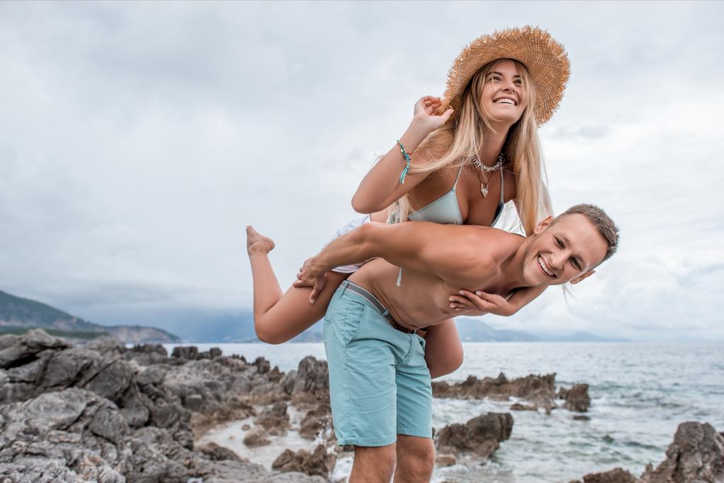 χαμηλή γωνία προβολής του happy νεαρό ζευγάρι piggybacking σχετικά με βραχώδη παραλία του Μαυροβουνίου - Φωτογραφία, εικόνα