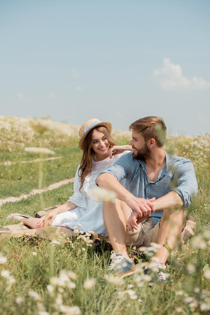 cheerful young lovers sitting on blanket in field with wild flowers on summer day - Photo, Image