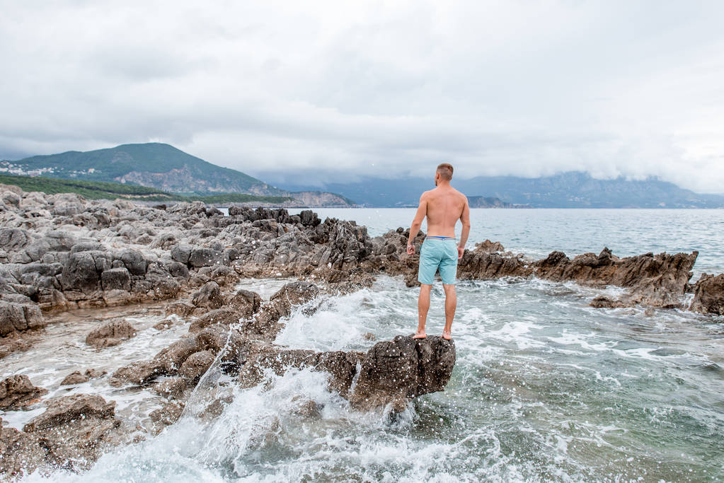 ハンサムな筋肉若い男岩のビーチの上に立って、モンテネグロの雄大な海の景色を見ての背面図 - 写真・画像