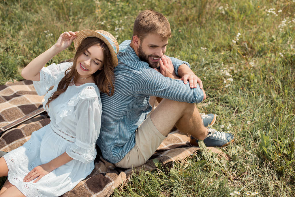 Hochwinkelaufnahme eines lächelnden Paares, das Rücken an Rücken auf einer Decke im Feld mit wilden Blumen an einem Sommertag ruht - Foto, Bild