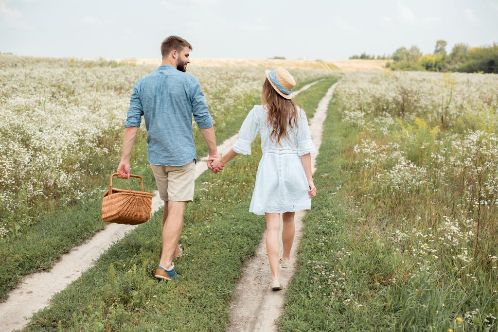 Rückansicht eines Paares mit Picknickkorb, das Händchen hält, während es im Feld geht - Foto, Bild