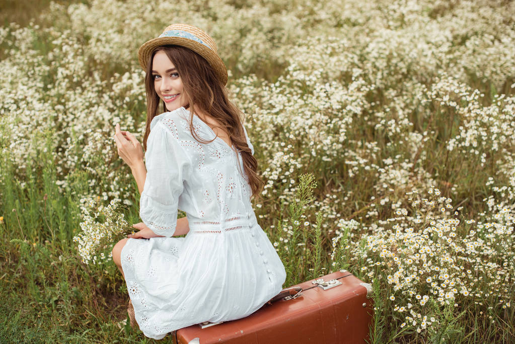 back view of smiling woman in white dress with bouquet of wild camomile flowers sitting on retro suitcase on meadow - Photo, Image