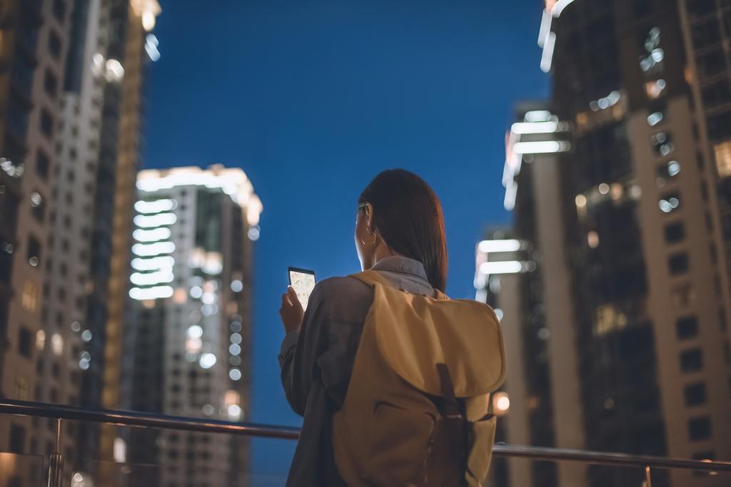 Hátulnézet nő a hátizsák és a smartphone-val megjelenítése a képernyőn kézzel állva éjszakai város utca - Fotó, kép