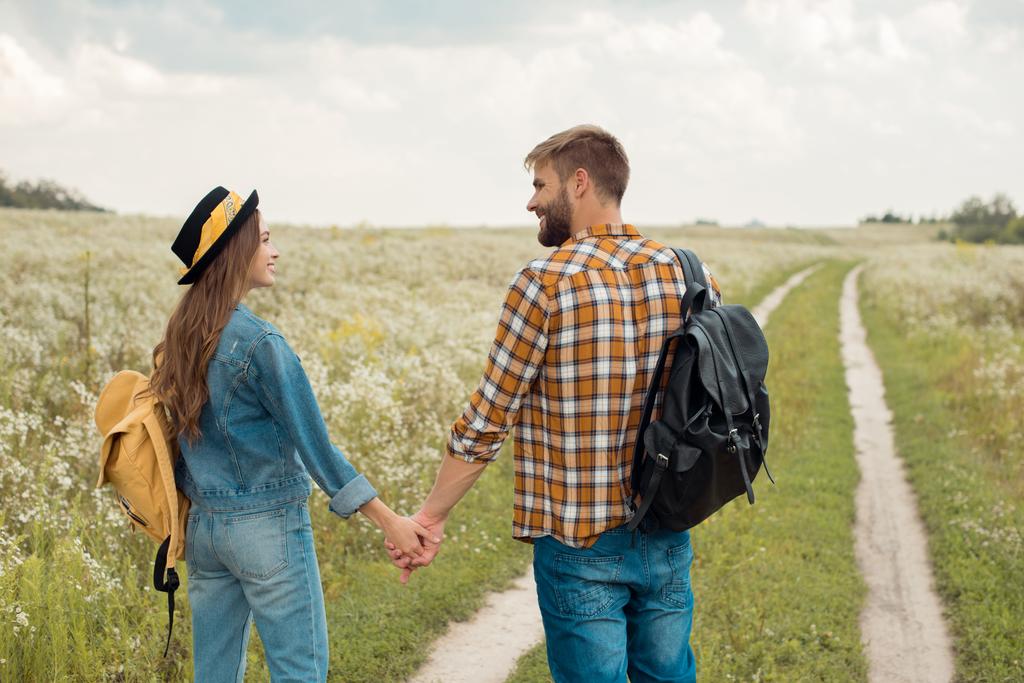 rear view of happy lovers with backpacks holding hands in field with wild flowers - Photo, Image