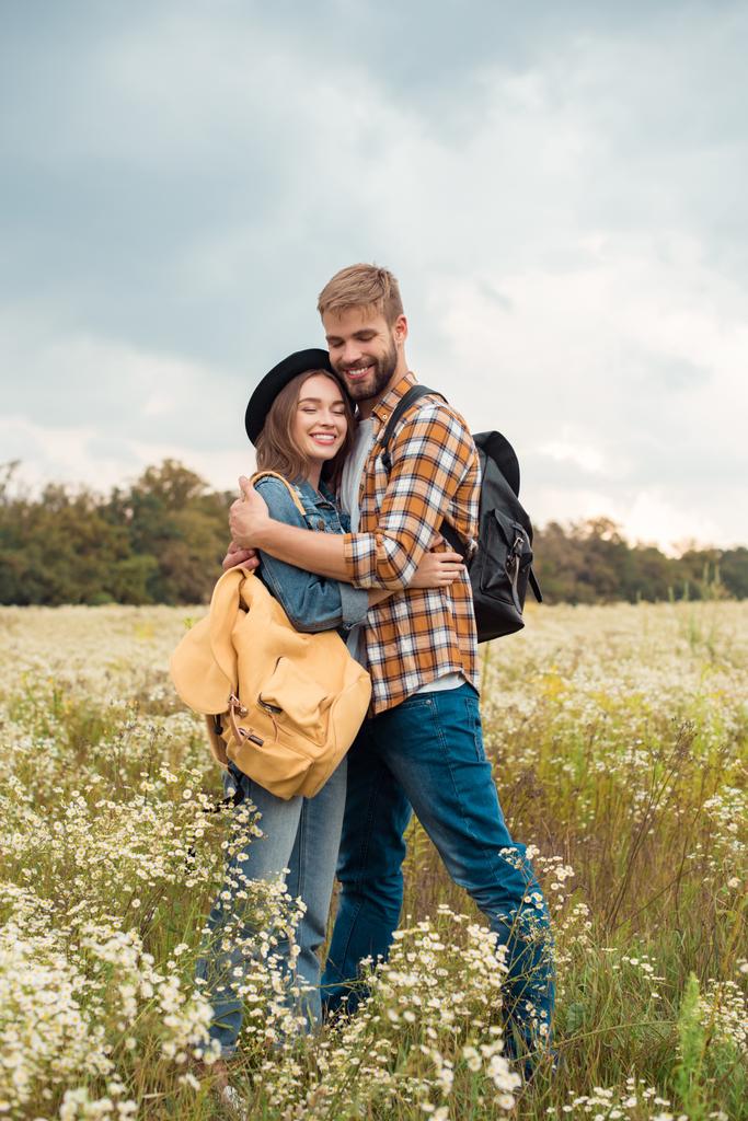 young couple with backpacks hugging in summer field with wild flowers - Photo, Image
