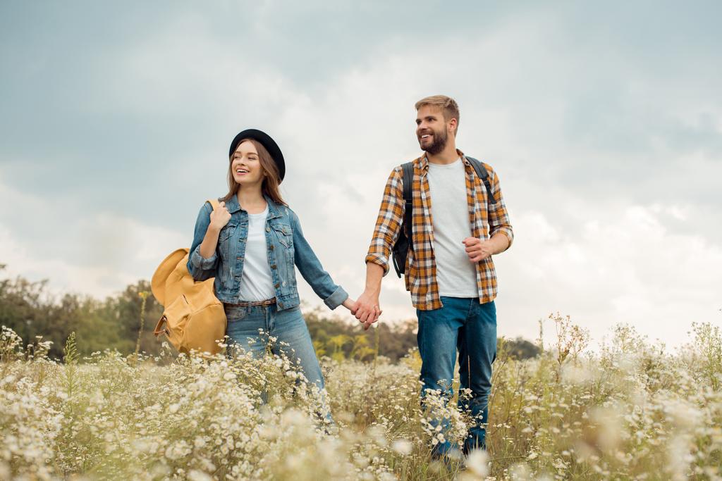 smiling couple with backpacks holding hands while walking in summer field with wild flowers - Photo, Image