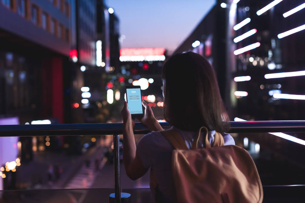 Widok kobiety z plecaka i smartphone z skype logo z powrotem na ekran w rękach stojąc na ulicy miasta w nocy - Zdjęcie, obraz