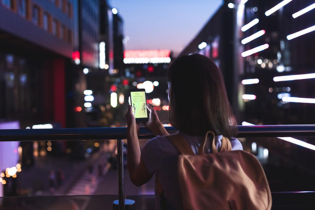 back view of woman with backpack and smartphone with shopping website on screen in hands standing on night city street - Photo, Image