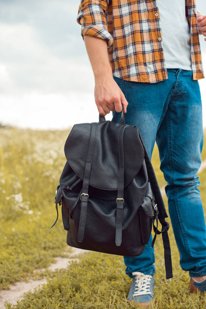 частичный вид человека в джинсах, держащего черный кожаный рюкзак в поле
 - Фото, изображение