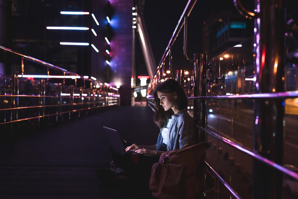 side view of young woman using laptop on street with night city lights on background - Photo, Image