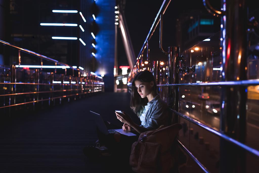 Μερική άποψη του νεαρή γυναίκα με φορητό υπολογιστή στα γόνατα χρησιμοποιώντας το smartphone με την πόλη το βράδυ στο φόντο - Φωτογραφία, εικόνα