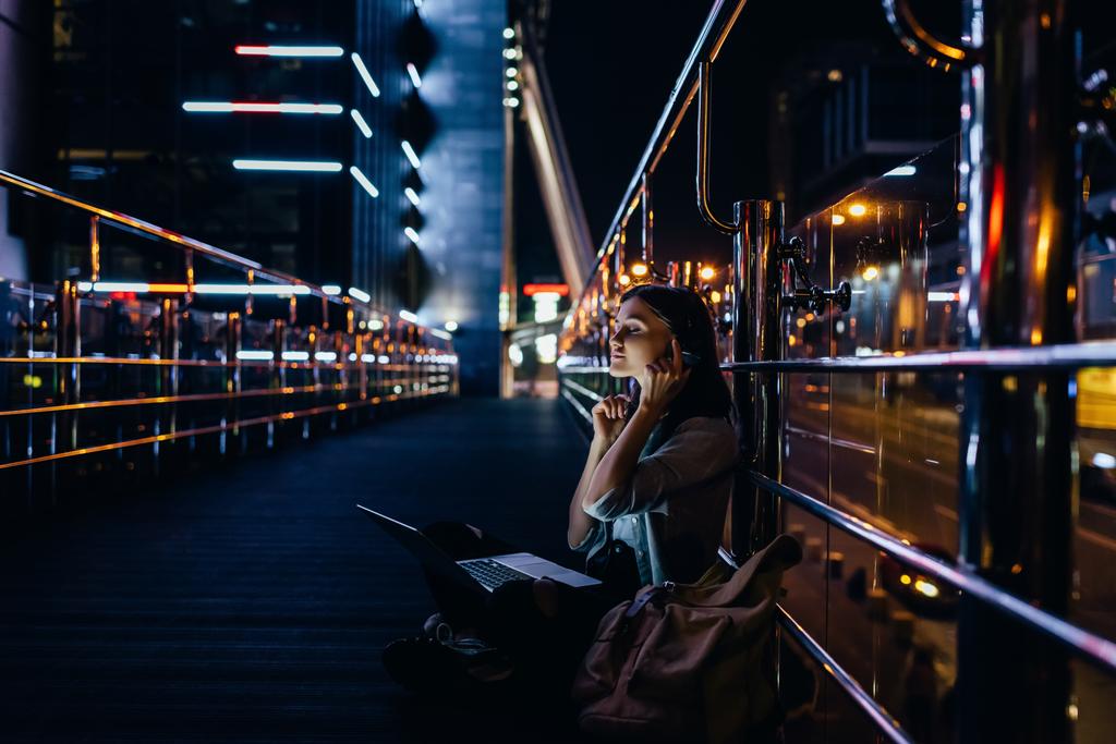 vista lateral da mulher ouvindo música em fones de ouvido com laptop de joelhos na rua da cidade à noite
 - Foto, Imagem