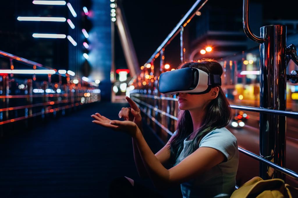 молода жінка в гарнітурі віртуальної реальності сидить на вулиці з нічним містом на фоні
 - Фото, зображення