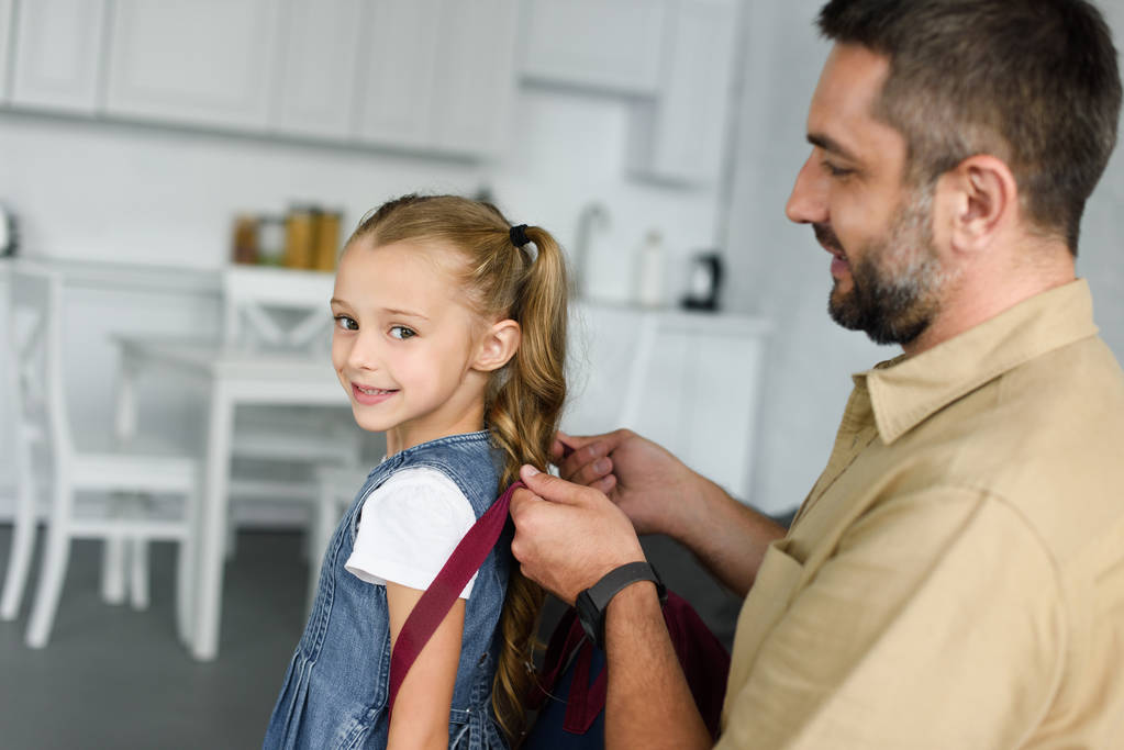 боковой вид отца, помогающего дочери носить рюкзак дома, обратно в школу
 - Фото, изображение