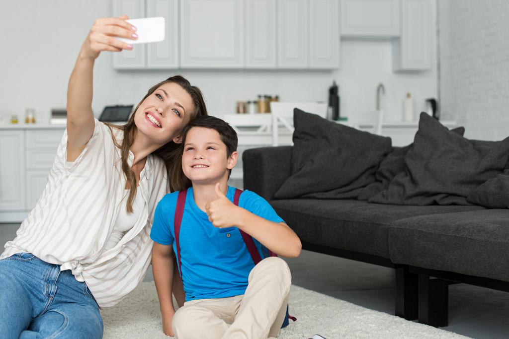 母と息子のバックパックと学校の最初の日に自宅でスマート フォンの selfie を一緒に取るを親指で笑みを浮かべてください。 - 写真・画像