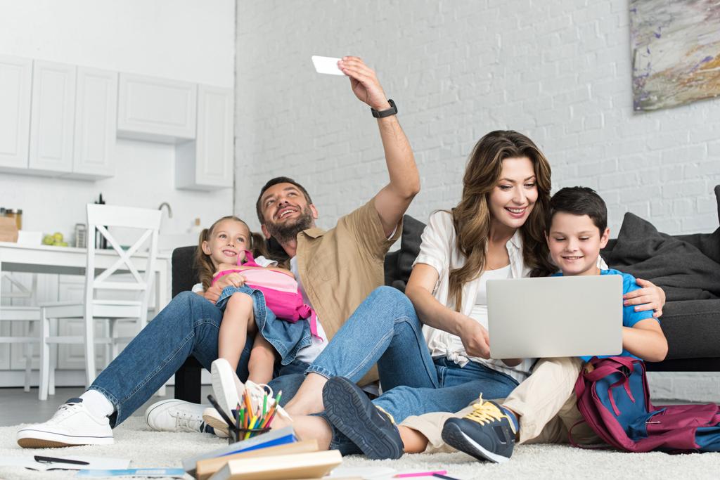 sonrientes padre e hija tomando selfie en el teléfono inteligente, mientras que la madre y el hijo usando el ordenador portátil juntos en casa
 - Foto, imagen