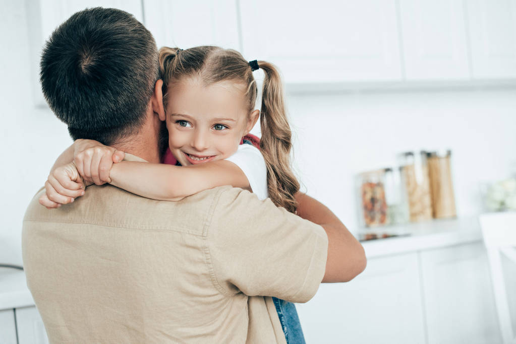 otec a usmívající se dcera s batohem objímaly v kuchyni doma, zpět do koncepce školy - Fotografie, Obrázek