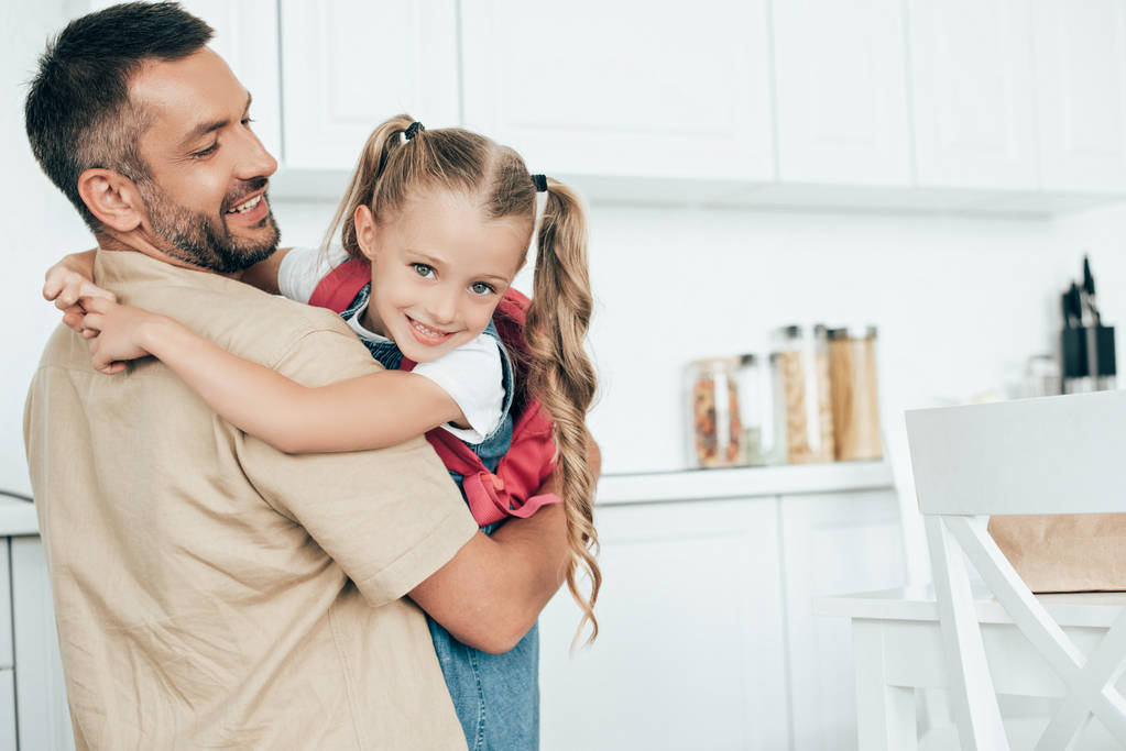 father and little daughter with backpack hugging each other in kitchen at home, back to school concept - Photo, Image