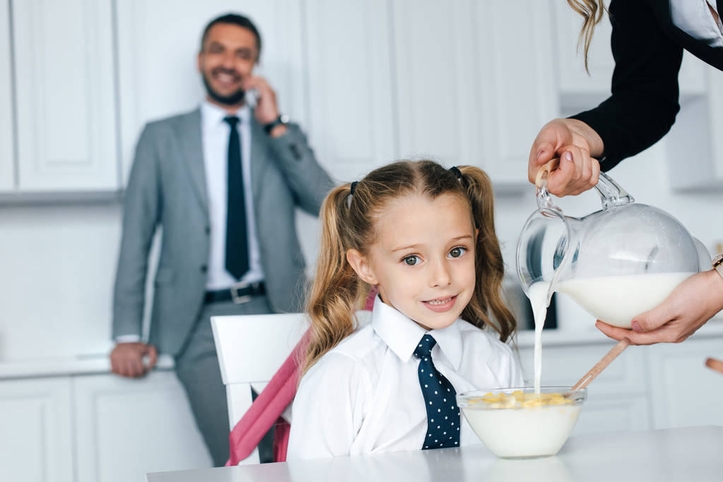 selective focus of kid in school uniform at table with breakfast and mother pouring milk into bowl with crunches at home, back to school concept - Photo, Image