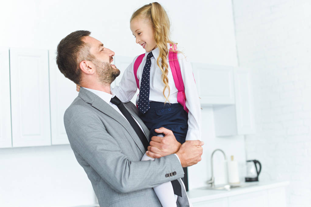 portrait of happy father in suit holding daughter in school uniform with backpack on hands in kitchen, back to school concept - Photo, Image
