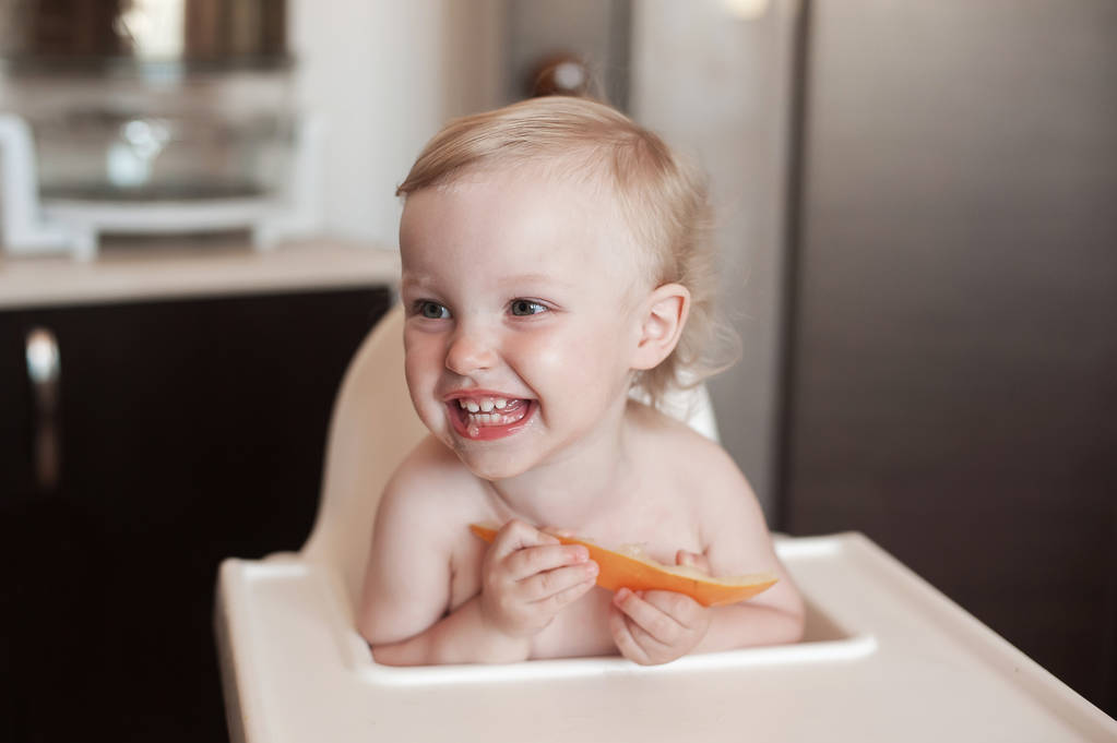 Малышка ест медовую дыню. Смешное смеющееся дитя на кухне. Веселый ребенок в кресле
 - Фото, изображение
