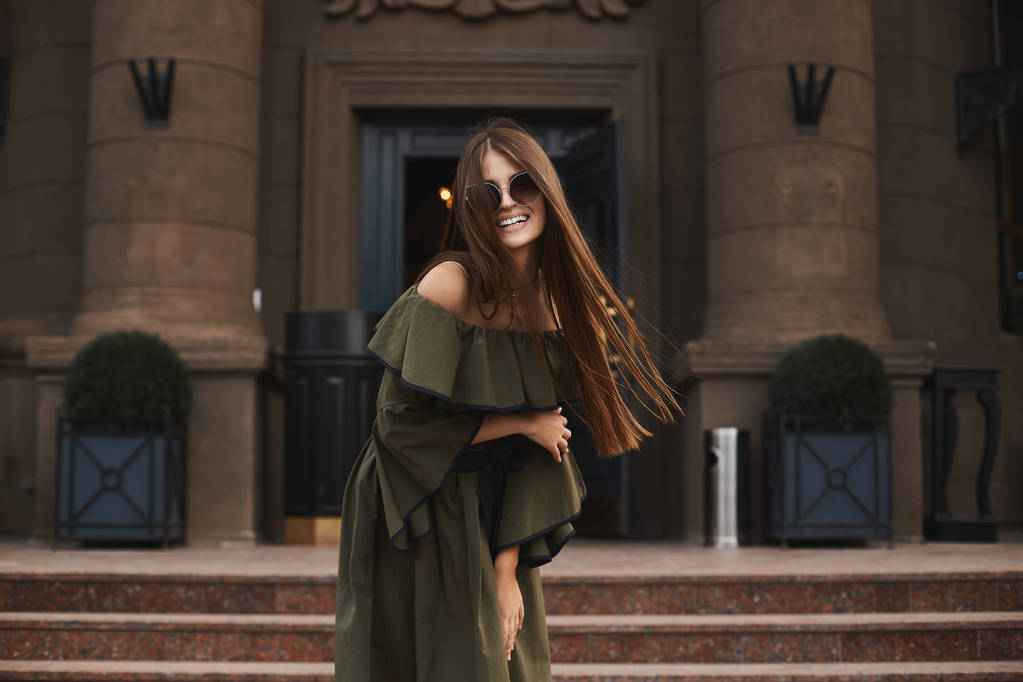 Piękne i modne model brunetka dziewczyna z czarujący uśmiech, w stylowy sukienka z nagie ramiona i modne okulary przeciwsłoneczne, pozują na zewnątrz przed starego budynku - Zdjęcie, obraz