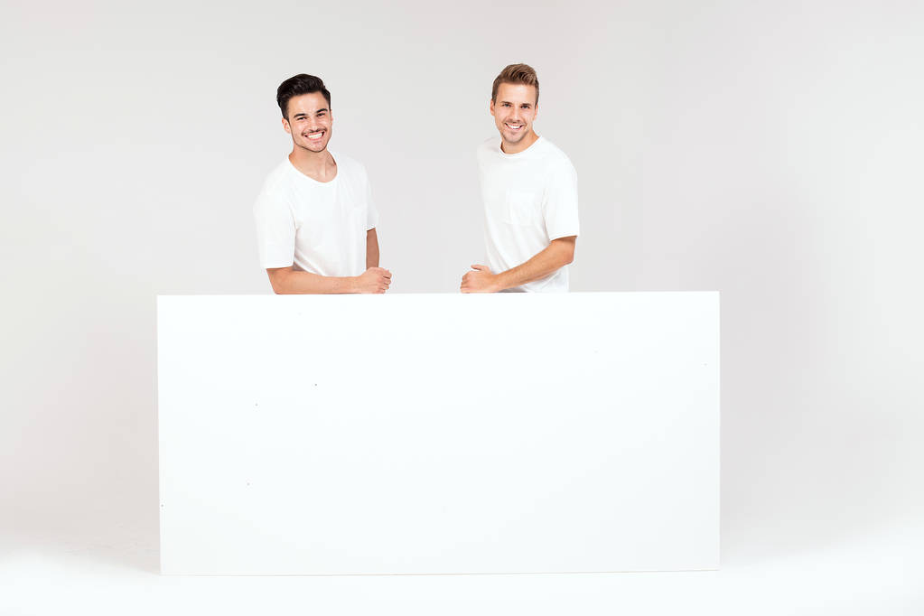 Dva mladí hezký kluci drží prázdný bílý nápis, vývěsní štít, izolované na bílém studio pozadí. - Fotografie, Obrázek