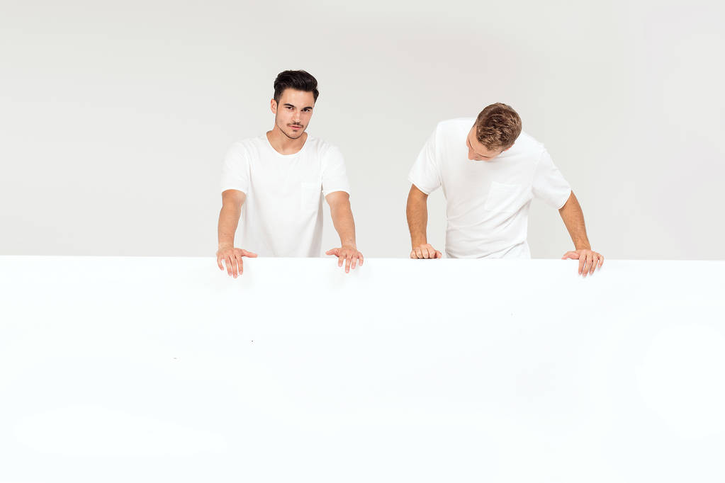 Двоє молодих красивих хлопців тримають порожній білий банер, вивіска, ізольовані на білому фоні студії
. - Фото, зображення