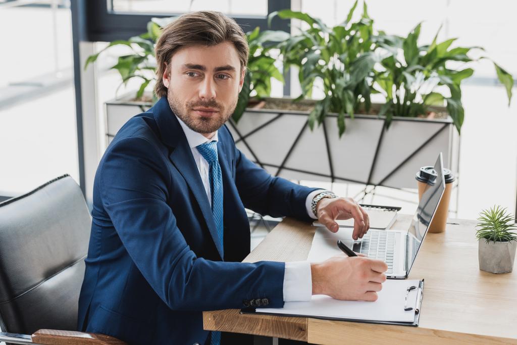 handsome young businessman looking away while writing on clipboard and using laptop in office - Photo, Image
