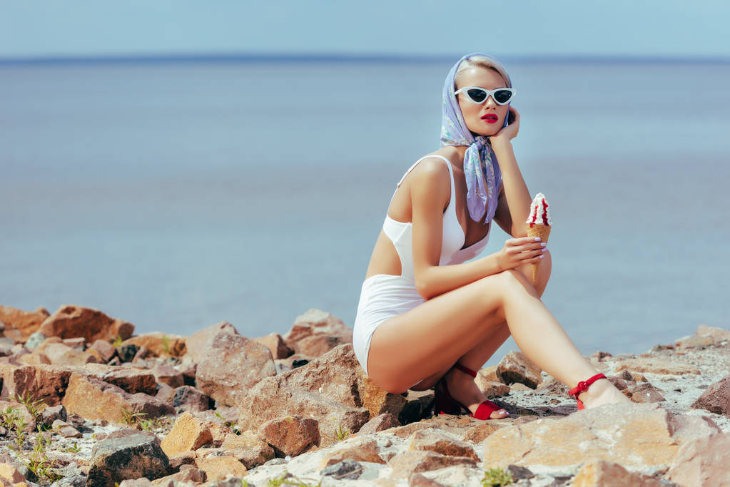 beautiful woman in vintage swimwear holding ice cream and posing on rocky beach - Photo, Image