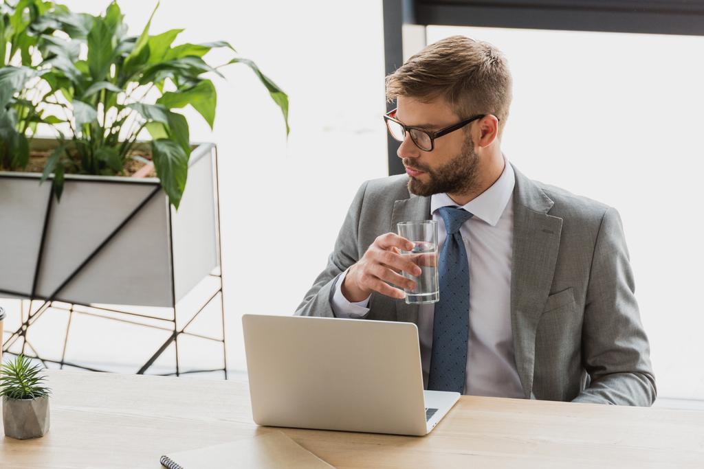 young businessman holding glass of water and looking away while using laptop in office - Photo, Image