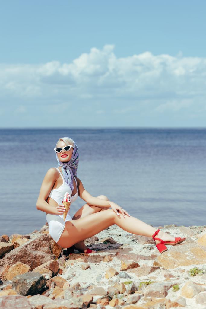 элегантная женщина в ретро купальнике держит мороженое и позирует на скалистом пляже в море
 - Фото, изображение