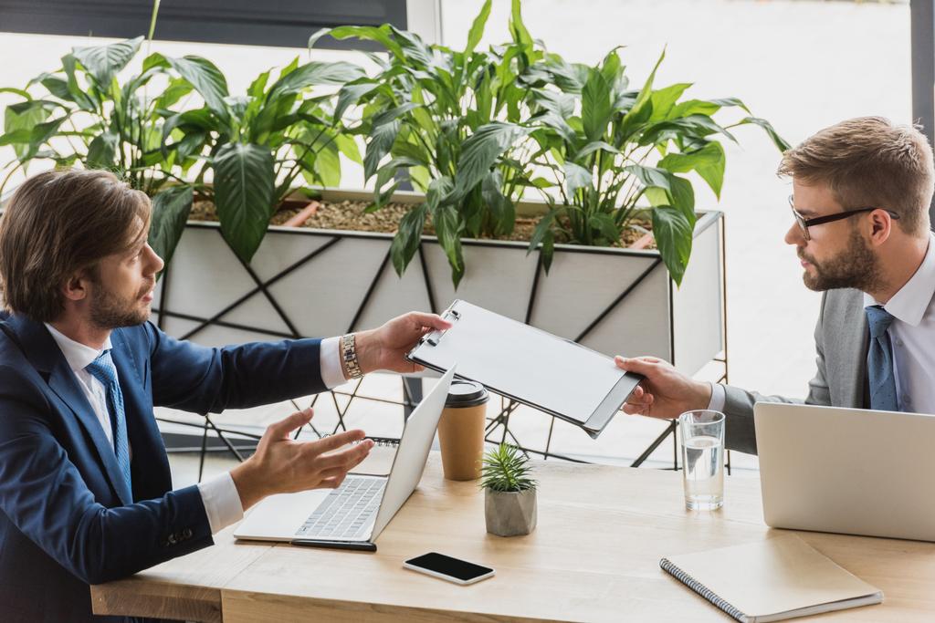 junge Geschäftsleute halten Klemmbretter in der Hand und schauen einander beim Arbeiten mit Laptops im Büro an - Foto, Bild