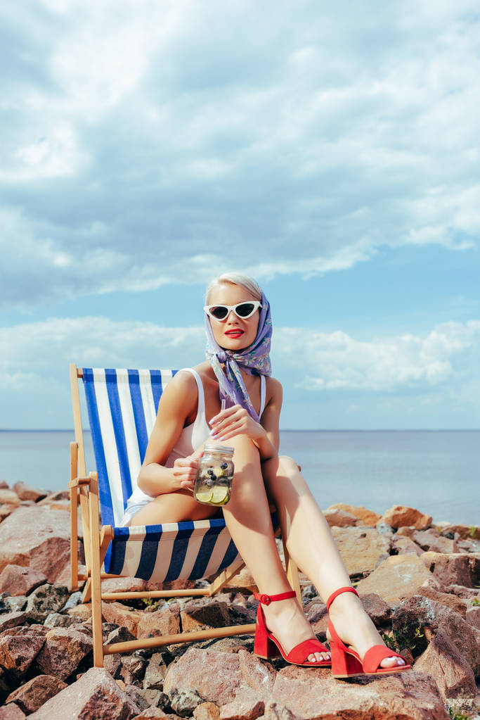 szép elegáns lány napszemüveg gazdaság jar, koktél, és pihentető a sziklás parton strand szék - Fotó, kép