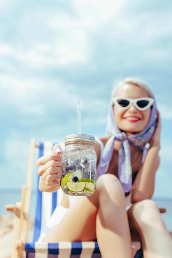 selective focus of smiling woman holding mason jar with lemonade and sitting in beach chair - Photo, Image