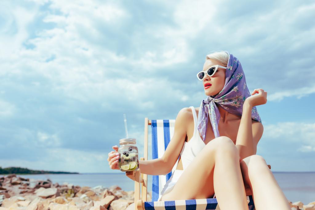 menina elegante atraente segurando limonada e relaxando em cadeira de praia na costa rochosa
 - Foto, Imagem