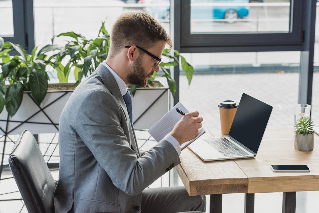 боковой вид бизнесмена, пишущего в блокноте и использующего ноутбук в офисе
 - Фото, изображение