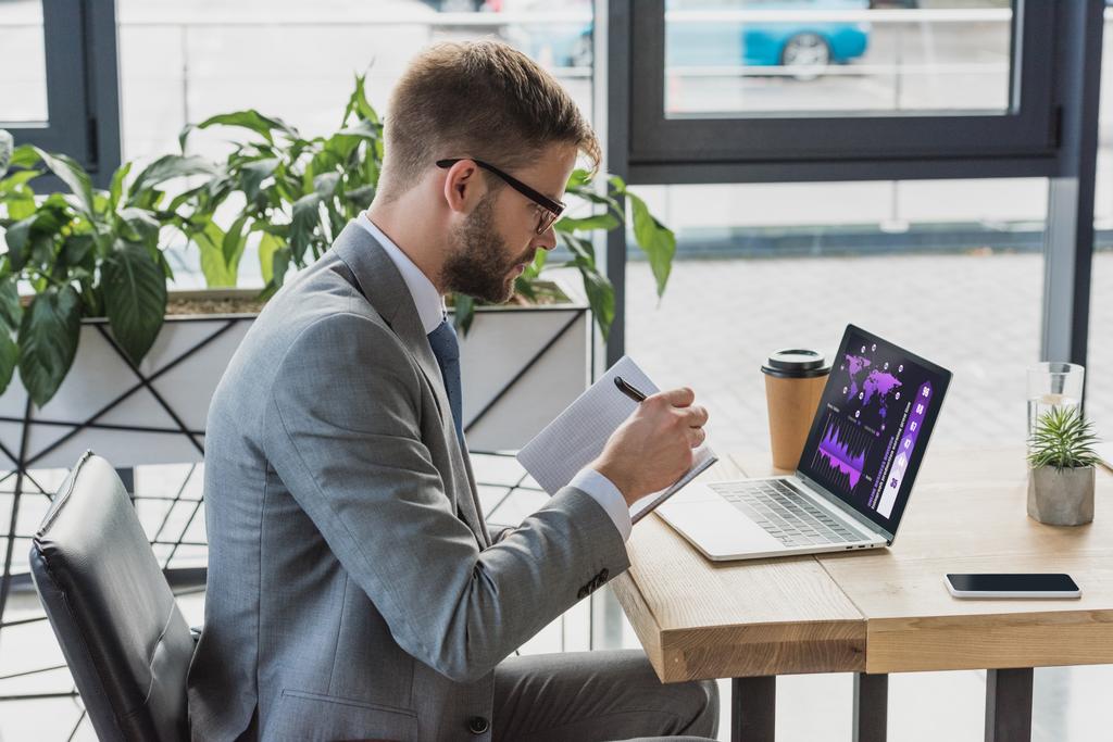 jeune homme d'affaires en costume et lunettes prenant des notes dans un carnet et utilisant un ordinateur portable avec des tableaux d'affaires
 - Photo, image