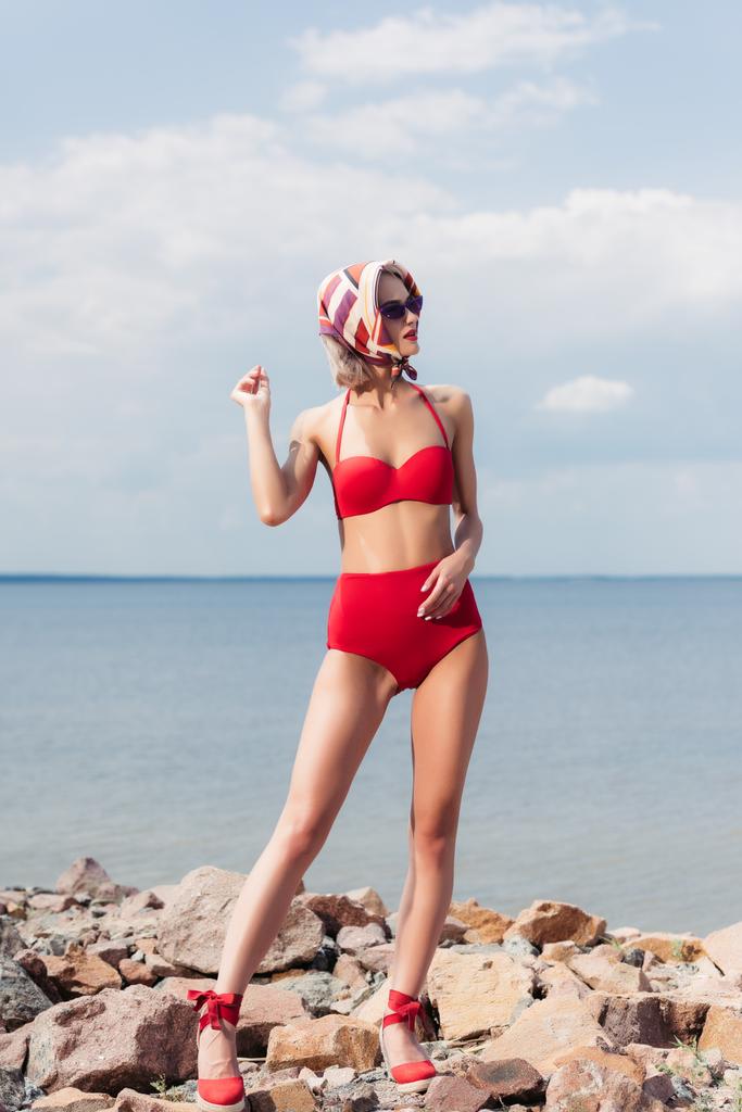 modelo elegante posando en bikini rojo y bufanda de seda en la playa rocosa cerca del mar
 - Foto, imagen