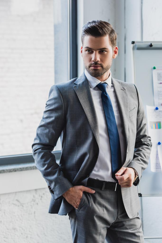 красивый серьезный молодой бизнесмен в костюме, стоящий с рукой в кармане в офисе
 - Фото, изображение
