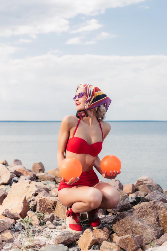 mujer elegante en bikini rojo retro, gafas de sol y bufanda de seda posando con bolas en la orilla rocosa
 - Foto, imagen