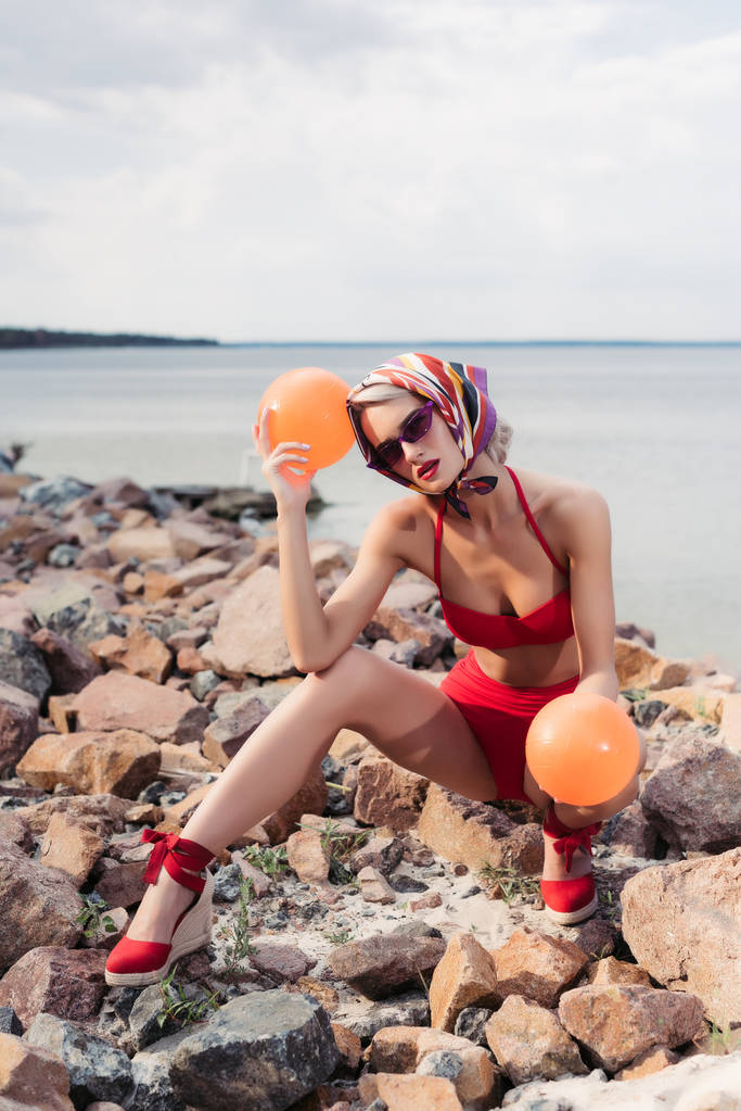 bela mulher elegante em biquíni vermelho, óculos de sol e cachecol de seda posando com bolas na costa rochosa
 - Foto, Imagem